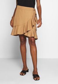 Tanfarvet omlægningnederdel med en flæsekant og bindelukning, lavet af blødt stof. Kombineret med sorte slip-on sko; modellen står foran en ensfarvet baggrund.