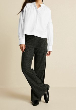 Osoba ubrana w białą bluzkę z długim rękawem, ciemnoszare spodnie o szerokich nogawkach oraz czarne mokasyny, stojąca ze skrzyżowanymi nogami na beżowym tle.