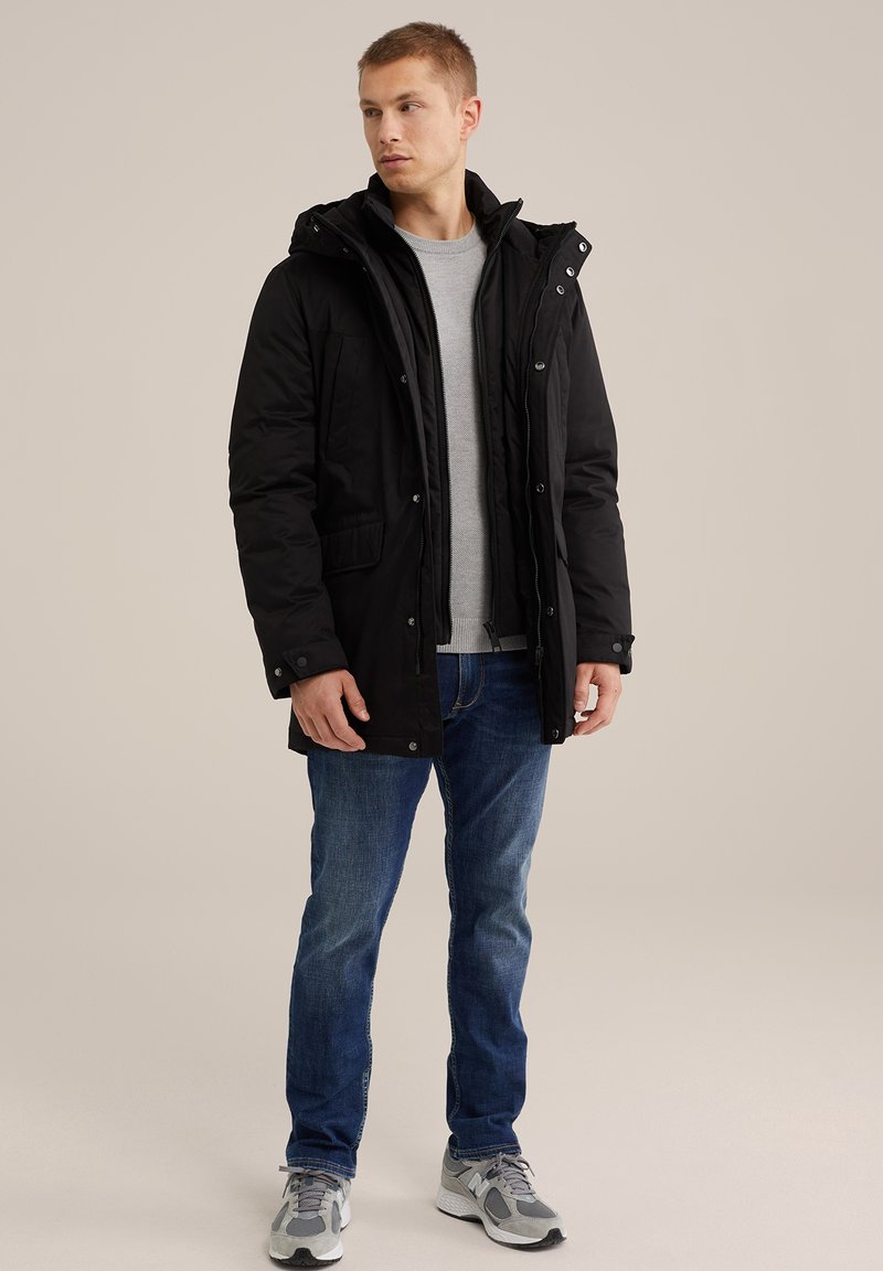 Schwarze isolierte Parka mit Kapuze, Reißverschluss, Druckknöpfen und vorderen Taschen. Getragen über einem grauen Pullover, kombiniert mit blauen Jeans und Turnschuhen.