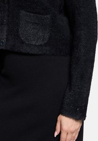 Czarny dziergany kardigan o puszystej fakturze, z naszywaną kieszenią z przodu i czarnymi guzikami, w połączeniu z dopasowaną czarną odzieżą poniżej.