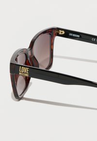 Suurte prilliraamidega kilpkonnakujulised prillid, millel on tumepruunid objektiivid ja mustal varrel kuldne „LOVE MOSCHINO“ kiri, moodsa ja massiivse disainiga.