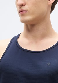 Μπλε ναυτικό τοπ από υφασμάτινη ύφανση, με στρογγυλή λαιμόκοψη και μικρό γκρι λογότυπο στο αριστερό στήθος.