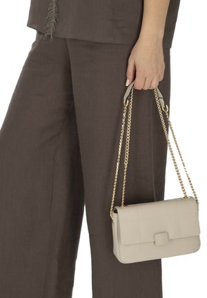 Ręka trzyma beżową skórzaną torebkę z złotym łańcuszkiem, nosi brązowe spodnie szerokie i dopasowany top.