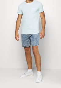 Hombre vestido con una camiseta de color azul claro con un bolsillo en el pecho, pantalones cortos de mezclilla con efecto desgastado, calcetines blancos y zapatillas blancas, de pie frente a un fondo blanco.
