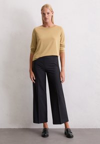 Senfgelbes Langarmoberteil kombiniert mit schwarzen weiten Hosen. Model trägt schwarze Loafers, die einen strukturierten Stoff und eine entspannte Passform zeigen.