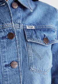 Svijetloplava traper jakna s bakrenim gumbima, Wrangler oznakom, džepom na prsima i zigzag detaljem u šivanju. Glatka tekstura i klasičan dizajn.