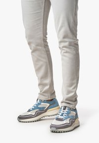 Sneakers con una tomaia in materiali misti blu e grigio, rete bianca testurizzata, suola leggera e lacci blu prominenti. Abbinati a pantaloni grigi chiari.