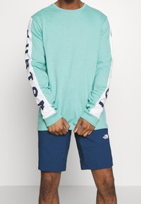 Mátově zelená košile s dlouhým rukávem a bílými rukávy, s námořnickým textem, doplněná námořnickými šortkami. Materiál vypadá lehce a prodyšně.