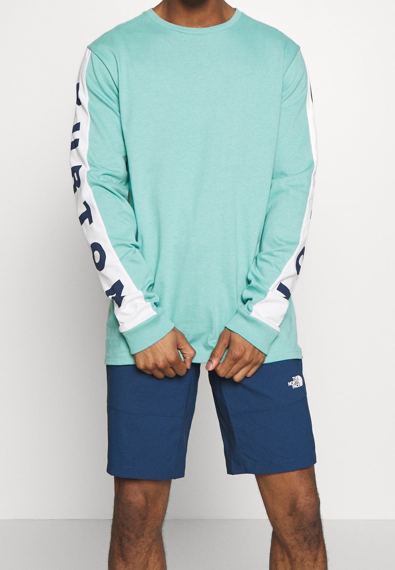 Mátově zelená košile s dlouhým rukávem a bílými rukávy, s námořnickým textem, doplněná námořnickými šortkami. Materiál vypadá lehce a prodyšně.