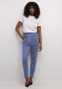 Lichtblauwe op maat gemaakte broek met een hoge taille en plooien, gecombineerd met een witte aansluitende t-shirt. Model staat met één hand in de zak.