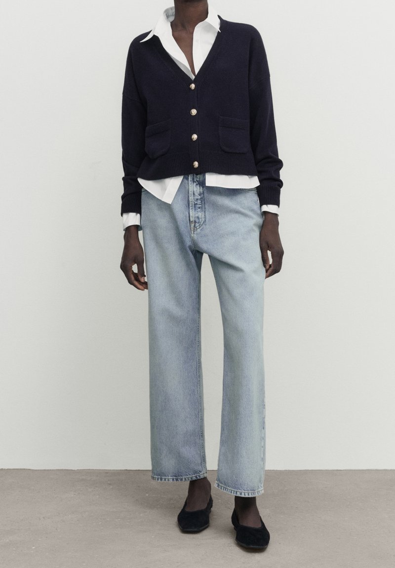 Personne portant un cardigan bleu marine boutonné sur une chemise blanche à col, un jean bleu clair à jambes larges et des chaussures plates noires, debout sur un sol en béton.