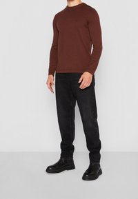 Pull long-sleeve marron foncé avec un col rond côtelé, associé à un jean noir et des bottines noires à semelles épaisses. Détail logo discret sur la poitrine.