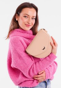 Beige zakulacená kabelka na rameno vyrobená z hladké kůže, s černým logem. Model má na sobě texturovaný růžový svetr.