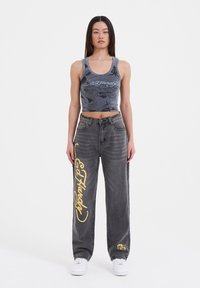 Gray crop top with dragon graphic, paired with loose gray jeans featuring yellow "Ed Hardy" text and pocket detail. White sneakers complete the look.