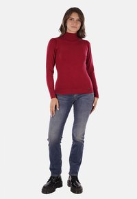 Maglione a collo alto rosso con un design a maglia testurizzata, abbinato a jeans blu e stivali neri con lacci, che mette in risalto una silhouette aderente.