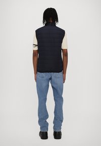 Marineblå puffervest med høy krage, quiltet design og en stilren finish, båret over en lys beige skjorte og kombinert med blå jeans.