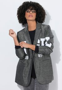 Frau mit lockigem Haar und schwarzer Brille passt die Ärmel eines grauen glitzernden Blazers mit weißem Text über einem schwarzen Hemd an.
