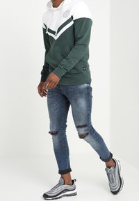 Sudadera con capucha verde y blanca con un diseño en forma de V, combinada con jeans de denim azul desgastados y zapatillas grises con acentos negros.