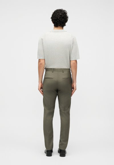 Άνδρας με σγουρά σκούρα μαλλιά, που φοράει ανοιχτό γκρι κοντομάνικο πουκάμισο και παντελόνι φόρμας ελιάς, στέκεται γυρισμένος προς τα πίσω σε απλό φόντο.