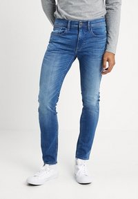 Man som bär slim-fit blå jeans, grå långärmad skjorta och vita sneakers, stående framför en enkel vit bakgrund.