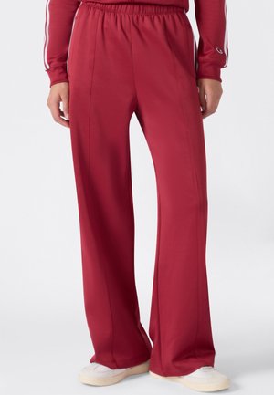 Personne portant un pantalon de survêtement rouge ample avec une ceinture élastique, associé à des baskets blanches, debout devant un fond uni.