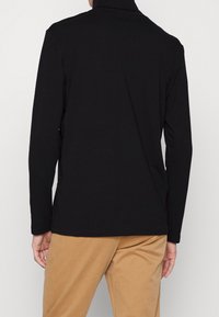 Mann in schwarzem Langarm-Turtleneck und beigen Hosen, von hinten vor einem schlichten hellen Hintergrund gezeigt.