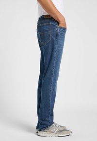 Jeans de mezclilla azul con un corte recto, dos bolsillos traseros, costuras visibles y una etiqueta de cuero marrón con la marca en la cinturilla.