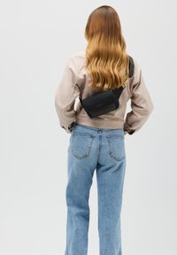 Svart axelremsväska i ett slätt material, rektangulär form, med ett guldfärgat axelband, som bärs över en ljusgrå jacka och blå jeans.