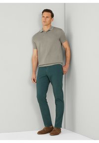 Man in grijs polo met korte mouwen, teal slim fit broek en bruine suede schoenen die tegen een lichtgrijze hoekmuur staat.