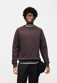 Μπλούζα μπορντό με στρογγυλή λαιμόκοψη και μανίκια ραγκλάν, φορεμένη πάνω από ένα ανοιχτό γκρι τοπ. Μαύρο καπέλο κρυμμένο στην τσέπη του παντελονιού.