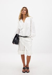 Balta mygtukų marškiniai su dviem krūtinės kišenėmis, derinami su baltais šortais su sukeltomis rankovėmis. Juoda rankinė, juodi sandalai su plokščiu padu.