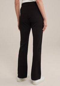 Černé rozšířené kalhoty s vysokým pasem, vyrobené z elastického materiálu, s hladkou texturou. Nosí se s bílou obuví.