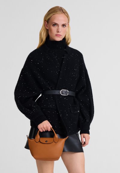 Black oversized knit sweater with silver speckles, high collar, and belt. Model holds a small tan leather bag. Black skirt visible.
