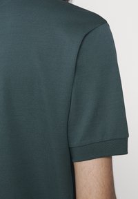 Gros plan sur le bras d'une personne portant un polo à manches courtes vert foncé en tissu texturé avec un poignet côtelé.