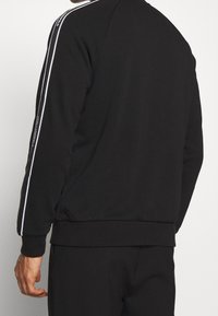 Felpa nera con strisce laterali bianche, polsini e orlo a coste, dotata di dettagli logo sulle maniche. Il materiale sembra morbido e liscio.