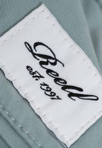 Label featuring the text "Beeld est. 1991" in black cursive on a white fabric tag, stitched onto a light blue-green textile surface.
