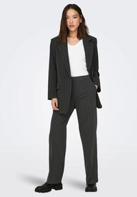 Giacca grigia a righe con ampie revers e pantaloni coordinati, abbinata a una camicia bianca a V e stivali con suola spessa nera.