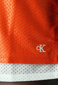 Oranžinė tinklinė medžiaga su mažomis skylutėmis, uždengta balta tinkline medžiaga. Apatinėje dešinėje kampe yra baltas išsiuvinėtas "cK" logotipas.
