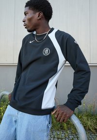 Sort og hvid Nike sweatshirt med rundt logo, lange ærmer, ribbede manchetter og en afslappet pasform. Bæres med lysblå denim jeans.