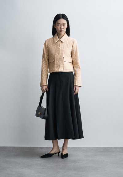 Femme portant une veste beige à carreaux, une longue jupe noire, des mules noires à talons, tenant un petit sac à main noir, debout devant un fond clair uni.