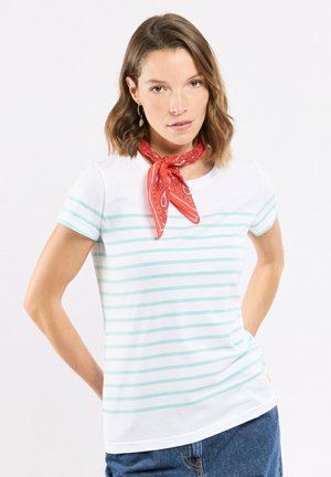Femme aux cheveux bruns mi-longs, portant des boucles d'oreilles dorées, un foulard rouge à motifs autour du cou, un t-shirt blanc à rayures bleu clair et un jean bleu.