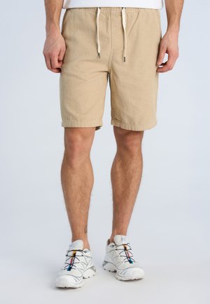 Mand iført beige kordfløjls-shorts med snøre og hvide atletiksko med flere farver på en lys baggrund.