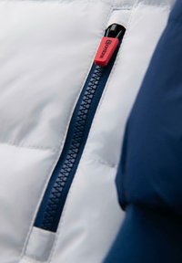 Biała kurtka puchowa z ciemnoniebieskim zamkiem błyskawicznym i czerwonym uchwytem. Miękka, gładka tekstura z pikowanym wzorem i akcentami szwów.