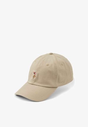 Lächelnde Frau mit langen blonden Haaren, die eine beige Kappe mit einem Polo-Spieler-Logo und eine schwarze Jacke mit Reißverschluss und passendem gesticktem Logo trägt.
