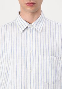 Homme portant une chemise blanche boutonnée avec de fines rayures verticales bleues et grises et une poche poitrine, col visible.