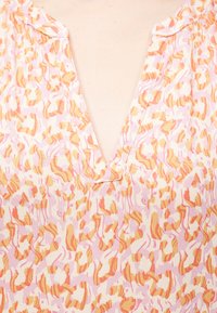 Μπλούζα με λαιμόκοψη σε σχήμα V, με ροζ, πορτοκαλί και λευκό αφηρημένο λουλουδάτο σχέδιο σε ανοιχτό ύφασμα, που δείχνει το πάνω μέρος του στήθους και την περιοχή του λαιμού.