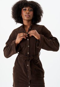 Brown corduroy shirt dress with long sleeves, button front closure, and a tied waist. Features a textured fabric and prominent buttons.