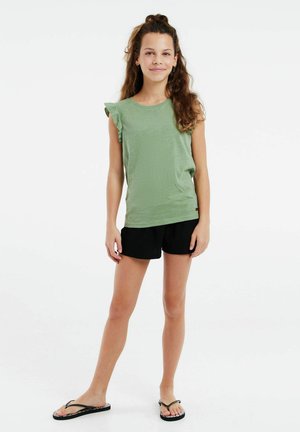 Top verde a maniche corte con dettagli arruffati sulle spalle, abbinato a pantaloni corti neri e ciabatte nere, in piedi su uno sfondo bianco uniforme.