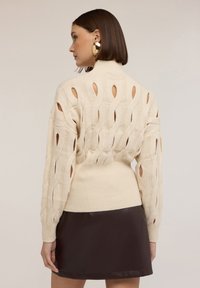 Beżowy sweter dzianinowy z wycięciami, wysoki kołnierz i ściągacz w pasie. Połączony z ciemnobrązową skórzaną spódnicą.