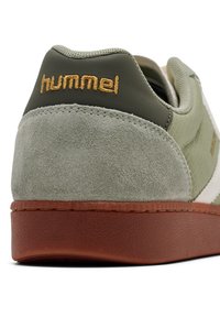 Groene suède en leren sneaker met een bruine rubberen buitenzool, voorzien van het "hummel"-logo in goud op de hiel en textuurpanelen.